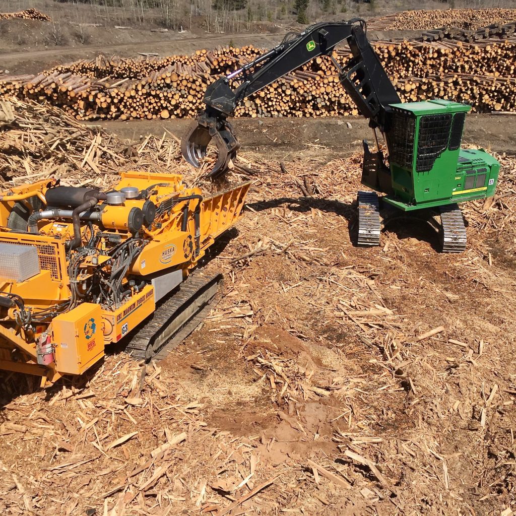 The yellow-orange wood grinder and wood waste recycler sits in the middle of piles of wood waste and chippings, and the green log loader loading wood into the grinder on a sunny day, operated by the Huska Holdings team.