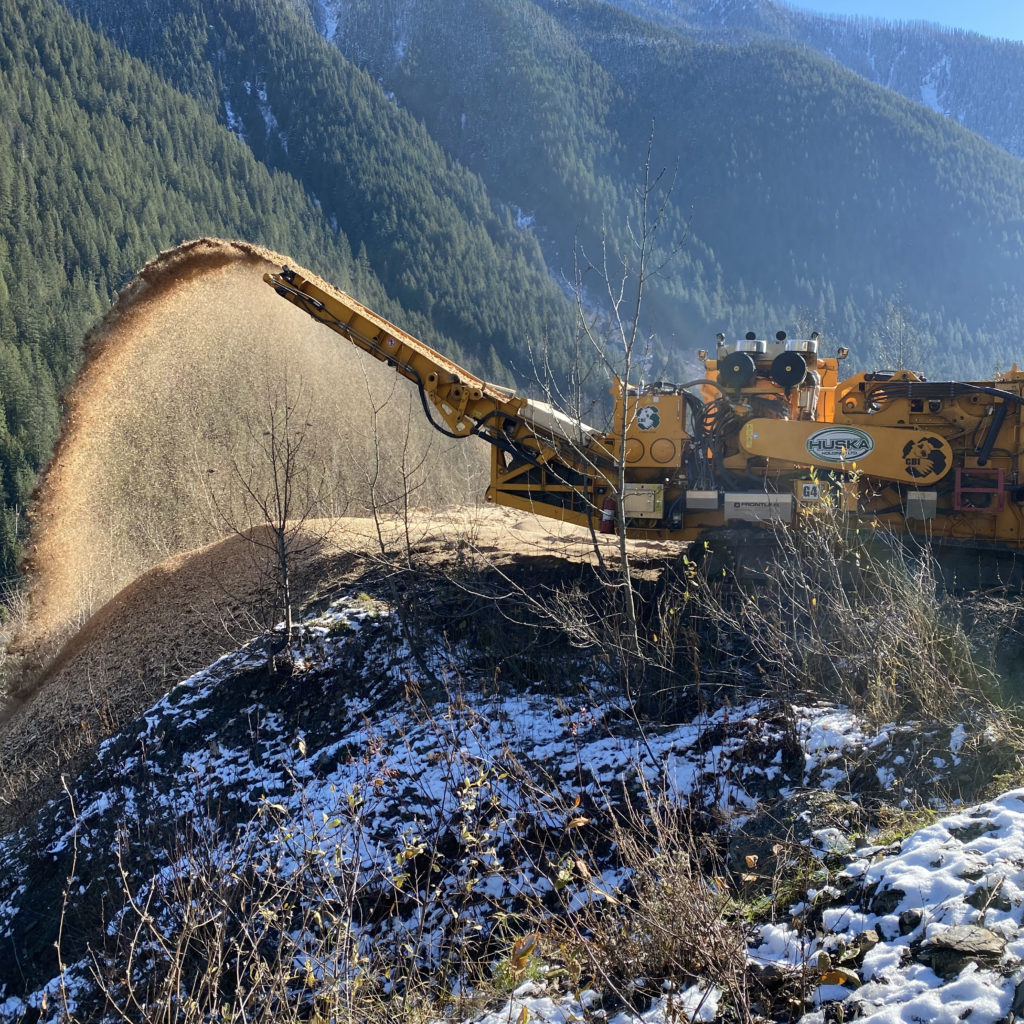 The yellow-orange wood grinder with the Huska Holdings logo shoots wood sawdust and chips over a hill, with patches of snow on the ground a a forest-covered mountain in the background on a sunny day, while the Huska Holdings team recycles wood waste.